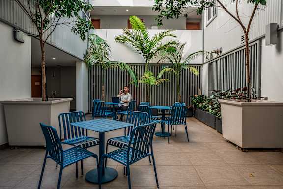 a man sitting at a table in a courtyard with blue chairs