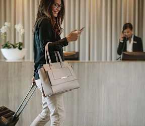 A woman with a suitcase is looking at her phone while standing in a room with a receptionist in the background. at Ovation at Town Madison, Madison
