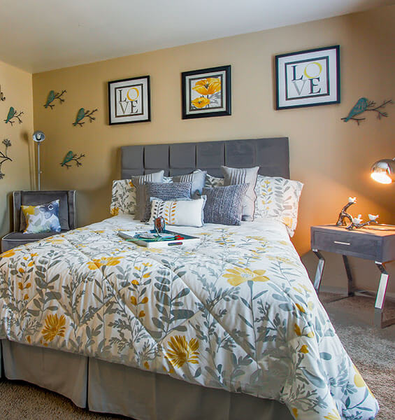 A bed with a floral comforter and matching pillows is in a room with a grey headboard and a desk with a lamp.