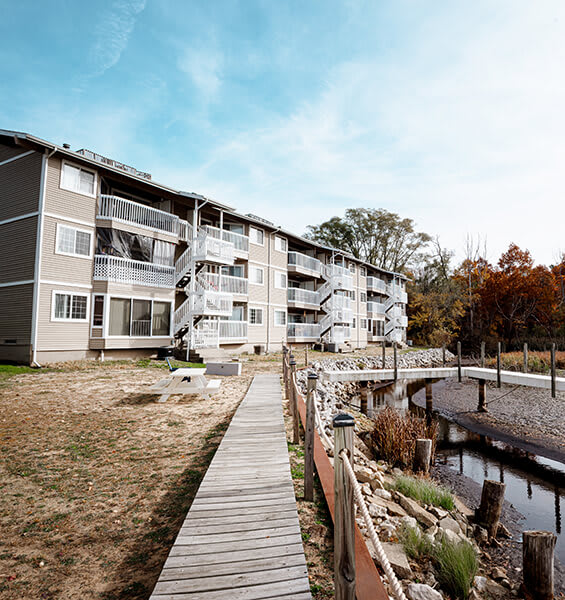 A wooden walkway leads to a building with a balcony and a small bridge over a body of water.