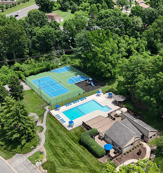 A tennis court is surrounded by a pool and a house.