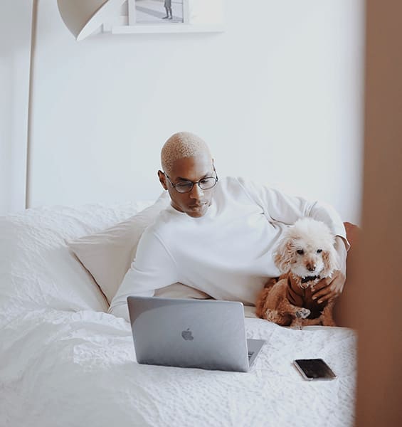 a man sitting on a bed with a dog and a laptop