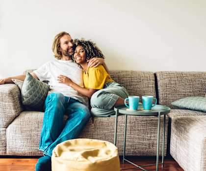 A couple is sitting on a couch with a potted plant to their left and a small table with cups on it to their right.