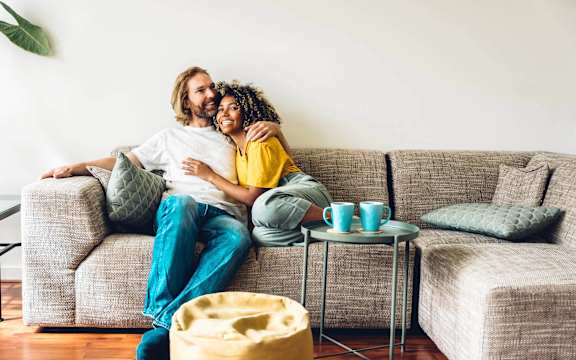 A couple is sitting on a couch with a potted plant to their left and a small table with cups on it to their right.