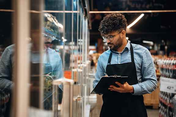 A man in a blue shirt and black apron is writing on a clipboard in a store.