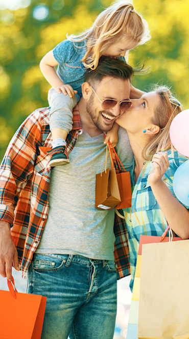 A family with a child on their shoulders, holding balloons and shopping bags.