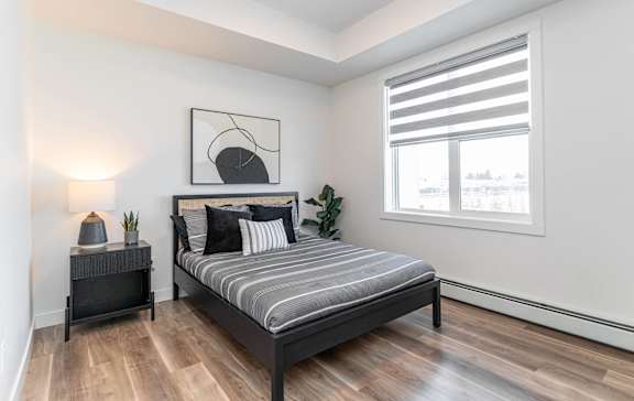 A bedroom with a bed, nightstand, and a window with blinds.at Vibe Apartments at Arbour Lake, Calgary Alberta