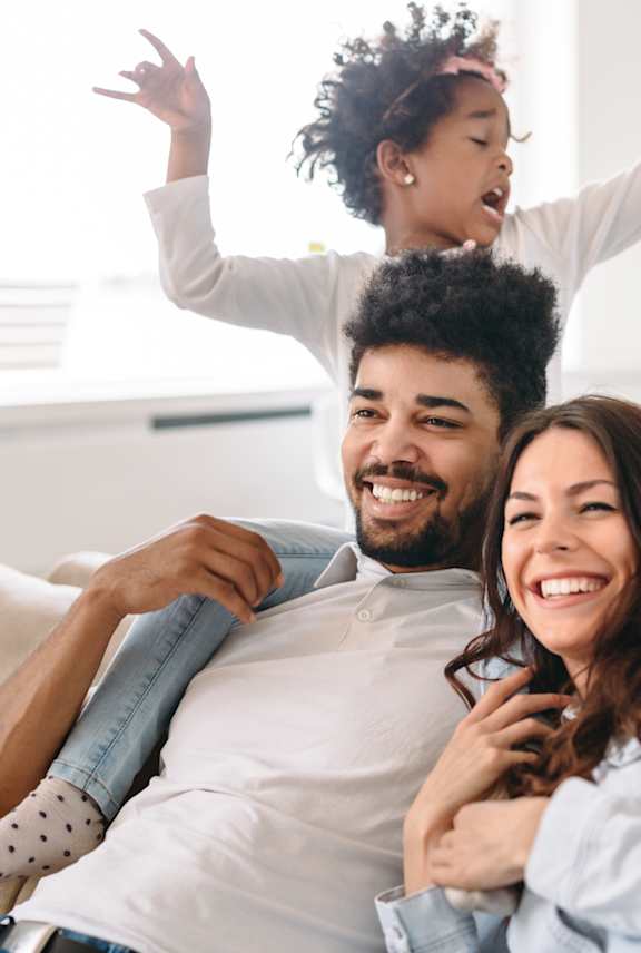 A young child is playfully standing on the shoulders of an adult while another adult sits on a couch.
