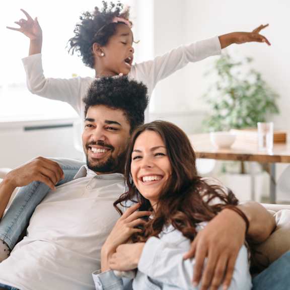 A young child is playfully standing on the shoulders of an adult while another adult sits on a couch.