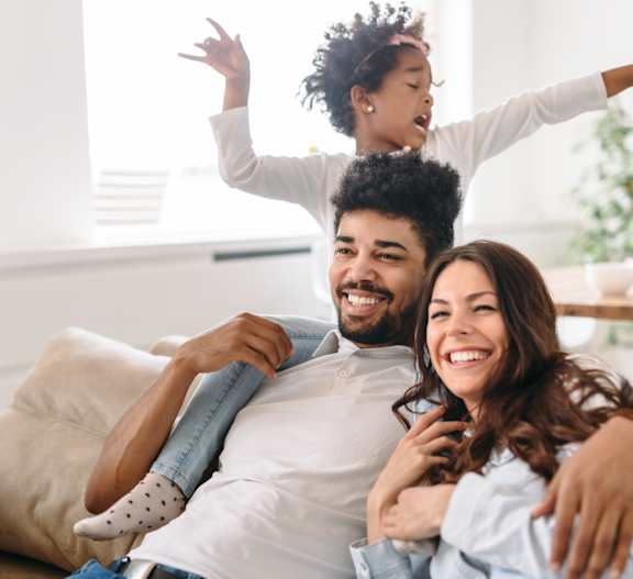 A young child is playfully standing on the shoulders of an adult while another adult sits on a couch.