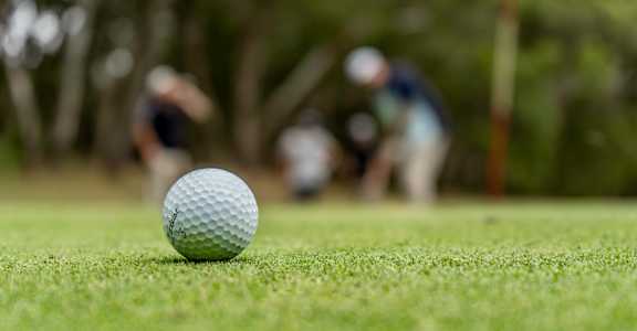 A golf ball sits on a green course with players in the background.