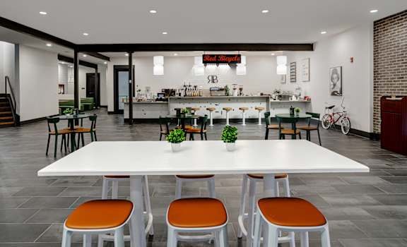 A white table with orange chairs is in the foreground of a room with a bar in the background.