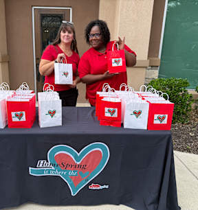 Two women in red shirts stand behind a table with bags that have a heart design on them.
