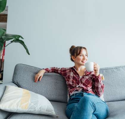 A woman is sitting on a grey couch holding a white mug.
