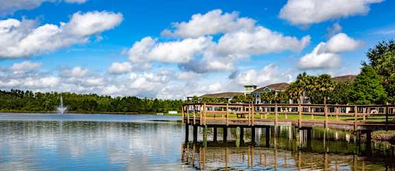 A wooden pier extends into a calm body of water with a fountain in the distance.