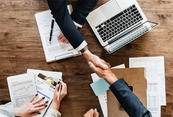 two people shaking hands over a table with laptops and papers