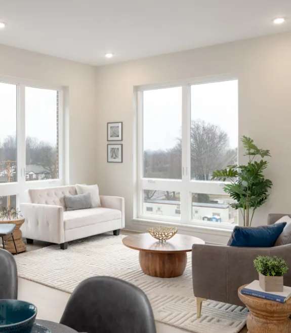 A living room with a white couch, a grey armchair, a wooden coffee table, and a large window with a view of the outside.