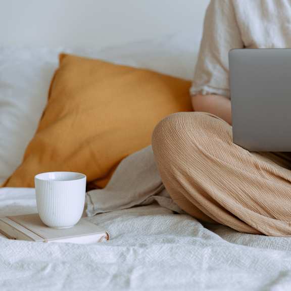 A person in a white shirt and beige pants is using a laptop on a bed with a white cup and a book.