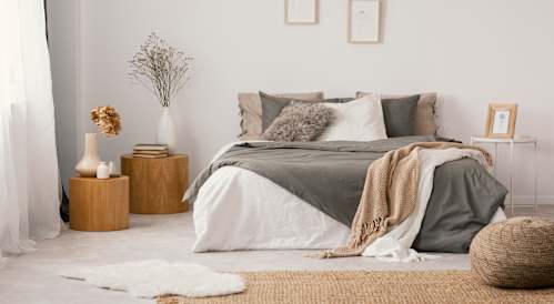 A bedroom with a bed covered in a grey blanket and pillows, a wicker basket on the floor, and a vase with dried flowers on a wooden stump.