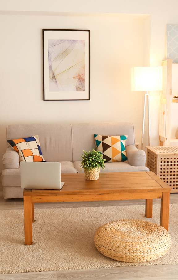 A living room with a grey sofa, a wooden coffee table, a wicker ottoman, a white stool, a wooden shelf, a white lamp, a white curtain, and a rug.