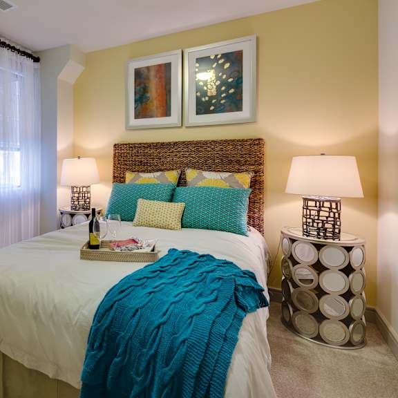 A bedroom with a large bed with a white bedspread and blue accents, and two lamps on top of bedside tables at Inspire Uptown Apartments.
