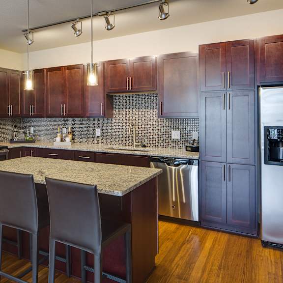 A kitchen with artwork, a light marble top counter, dark wood cabinets, refrigerator, and barstools at Inspire Uptown Apartments.