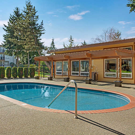 The large, turquoise outdoor pool with the pool house, bushes, and trees with the apartments in the background at Soundview Apartment Homes.