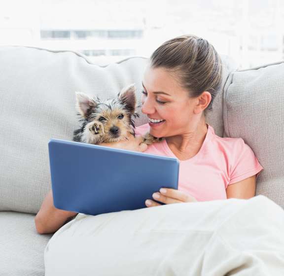 A woman is sitting on a couch with a dog and using a tablet.