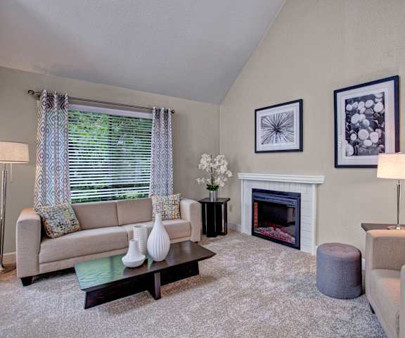 The spacious, neutral living room with a couch, throw pillows, large windows, a ceiling fan, wall art, plush chair, coffee table, decorations, a fireplace, large windows, and a TV at the Cove Apartment Homes. 