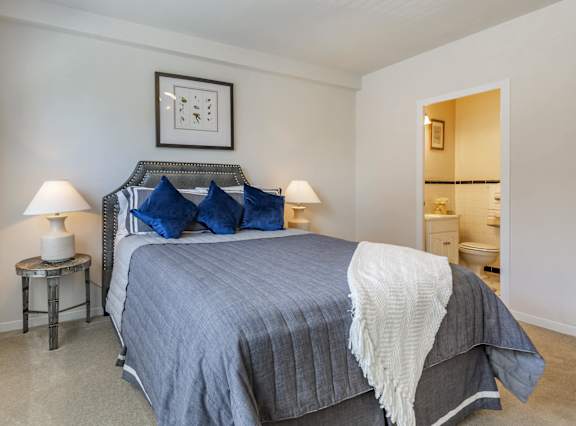 A bed with a grey comforter and a white blanket is in the foreground of a room with a framed picture on the wall.