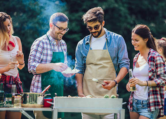 A group of people are gathered around a table, with one person wearing an apron and cooking food on a grill.