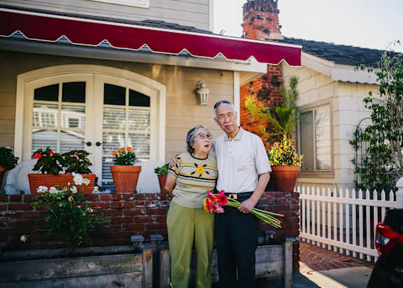 An elderly couple stand outside their house holding flowers.