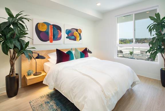 A bedroom with a large bed, a nightstand with a lamp, and two potted plants.