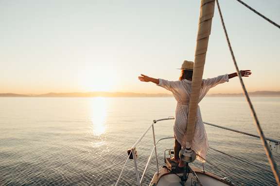A person on a sailboat with arms outstretched at sunset.