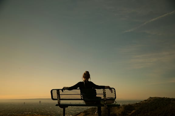 A person is sitting on a bench overlooking a cityscape at sunset.