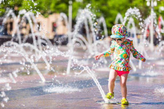 A little girl in a colorful outfit is playing in a water fountain.