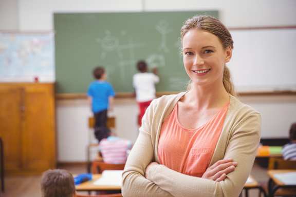 A woman teacher stands in front of a blackboard in a classroom with students in the background.