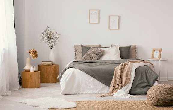 A bedroom with a bed covered in a grey blanket and pillows, a wicker basket on the floor, and a vase with dried flowers on a wooden stump.