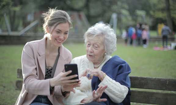 A young woman is showing something on her phone to an elderly woman on a park bench.