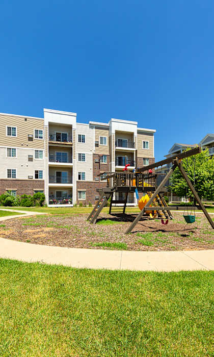 A playground with a swing set is in the middle of a grassy area in front of apartment buildings.