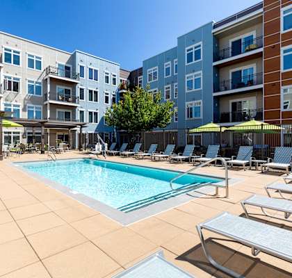 A pool surrounded by sun loungers and umbrellas in front of apartment buildings.