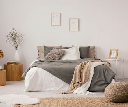 A bedroom with a bed covered in a grey blanket and pillows, a wicker basket on the floor, and a vase with dried flowers on a wooden stump.