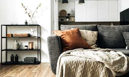 A grey couch with a blanket and pillows in a room with a shelf and decorations.