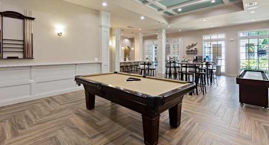 A pool table is in the middle of a room with wood flooring and a bar area.