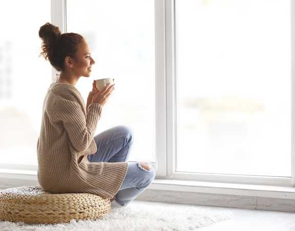 A woman in a beige sweater is sitting on a woven stool by a window.