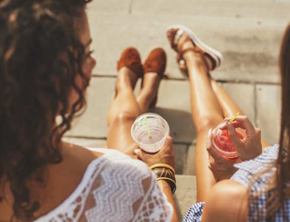 Two women sitting on the ground with their feet up, one holding a drink and the other holding a snack.