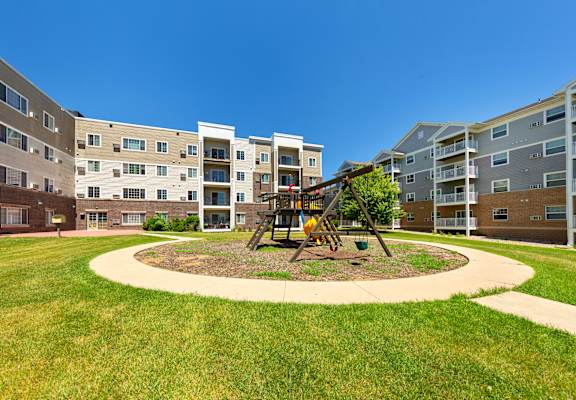 A playground with a swing set is in the middle of a grassy area in front of apartment buildings.