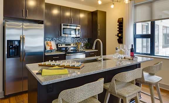 A kitchen with stainless steel appliances, marble countertops, a wine bottle, wine glasses, barstools, and large, dark cabinets at Inspire Apartments. 