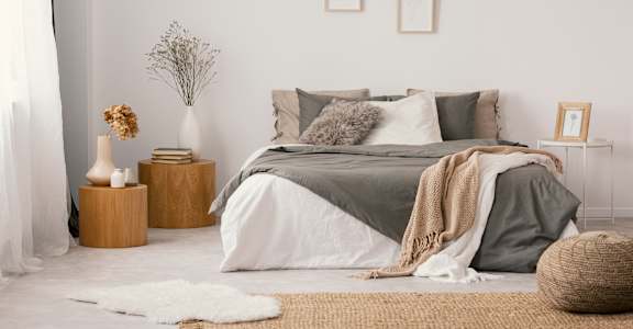 A bedroom with a bed covered in a grey blanket and pillows, a wicker basket on the floor, and a vase with dried flowers on a wooden stump.