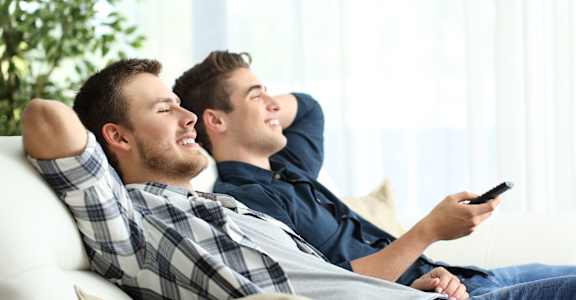 Two men are relaxing on a couch, one holding a remote control.